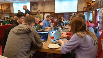 The image depicts a group of people seated around a table, working on laptops. They appear to be engaged in a collaborative activity or discussion, possibly related to software development or IT work, given the presence of code and screens open with what looks like GitHub repositories. The environment suggests they might be in an office setting, as indicated by the formal attire of some individuals and the professional-looking setup with laptops on a table.